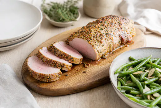 Filet mignon de porc entier doré à la croûte de moutarde et d’herbes, tranché en rondelles roses sur une planche en bois avec des haricots verts