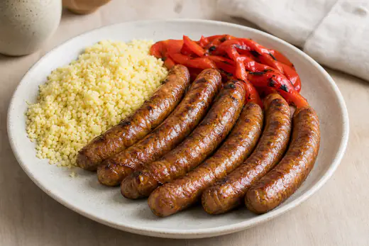 Six merguez bien dorées dans leur boyau, servies dans une assiette avec de la semoule et des poivrons grillés